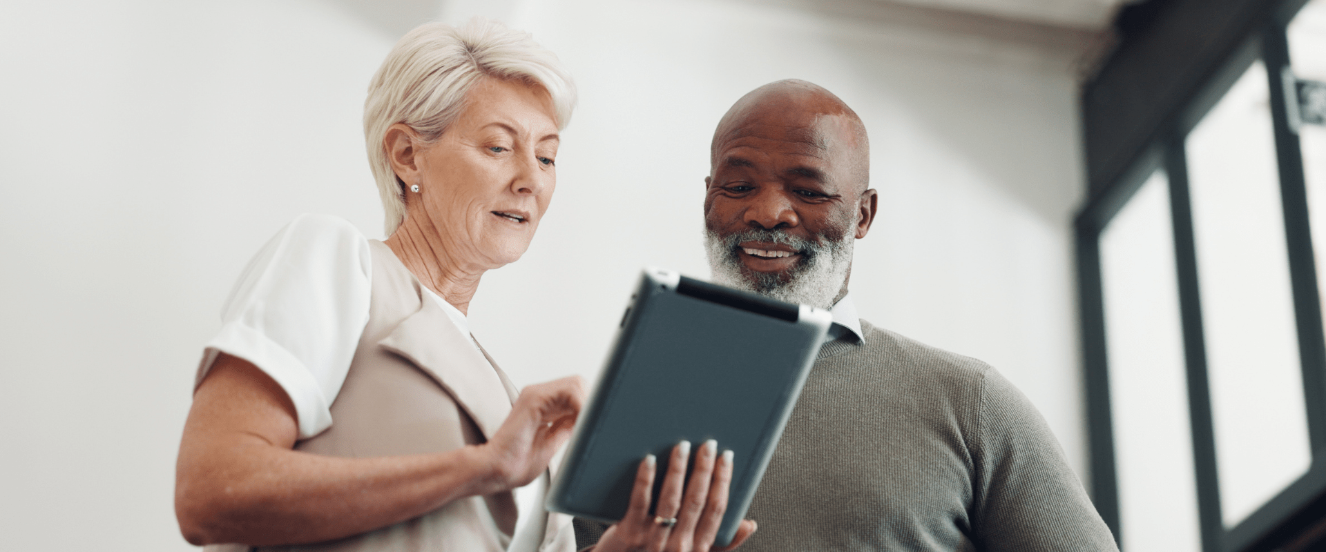 Two older workers stand in an office looking at a tablet. Two older workers stand in an office looking at a tablet.