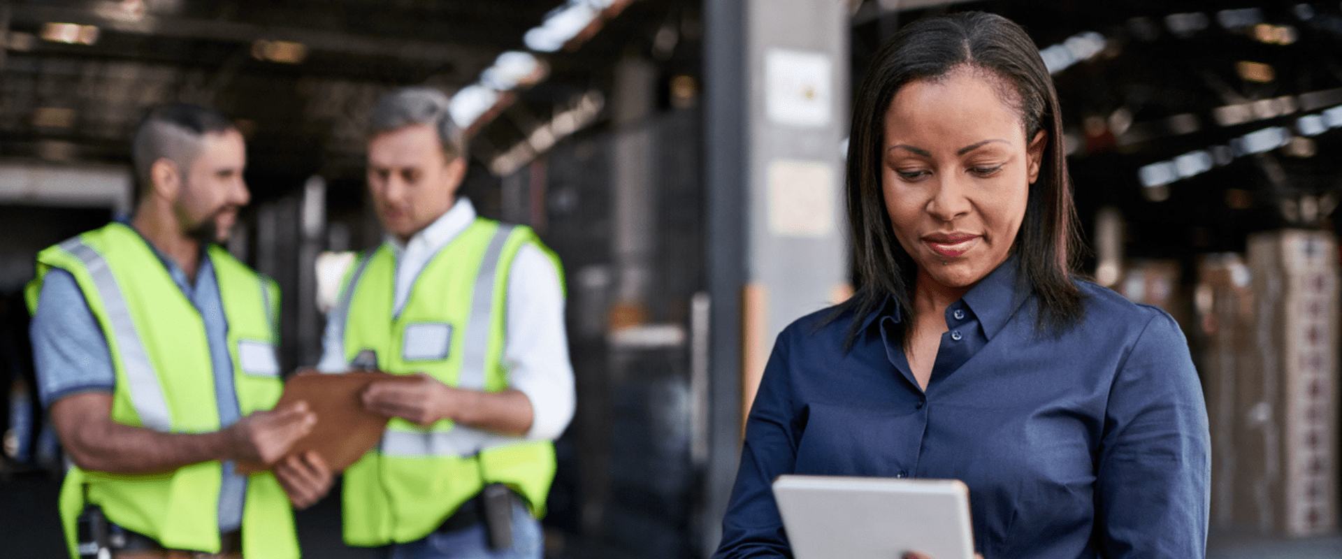 A worker looks at a tablet, while two others in safety vests talk behind them.