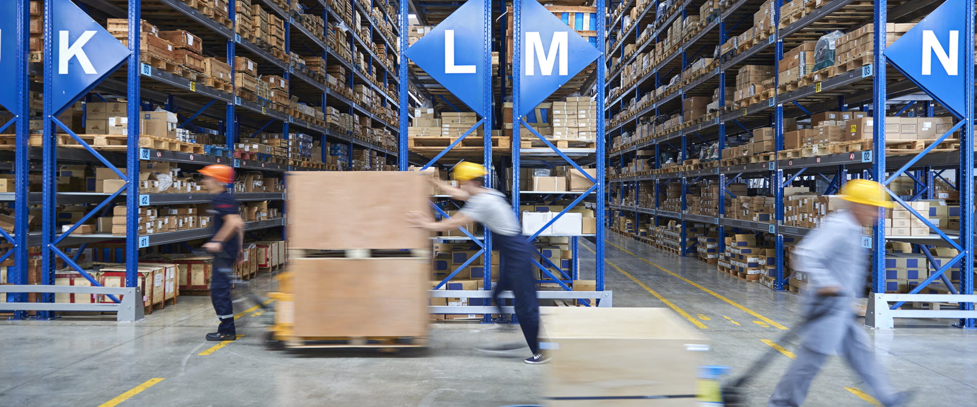 Workers briskly walk across the floor of a warehouse filled with floor-to-ceiling shelves, filled with boxes Workers briskly walk across the floor of a warehouse filled with floor-to-ceiling shelves, filled with boxes