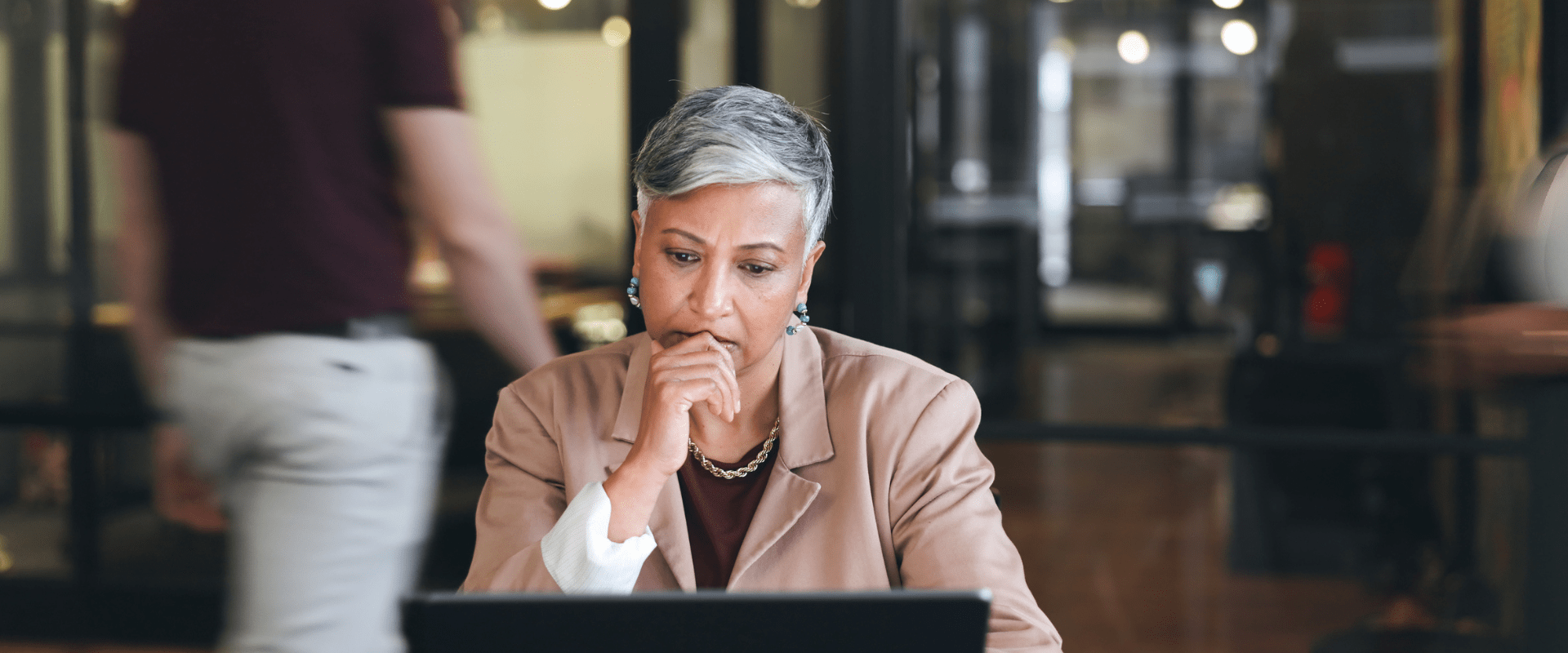 A manager sits at a laptop looking stressed, someone is walking out of the office behind them. A manager sits at a laptop looking stressed, someone is walking out of the office behind them.