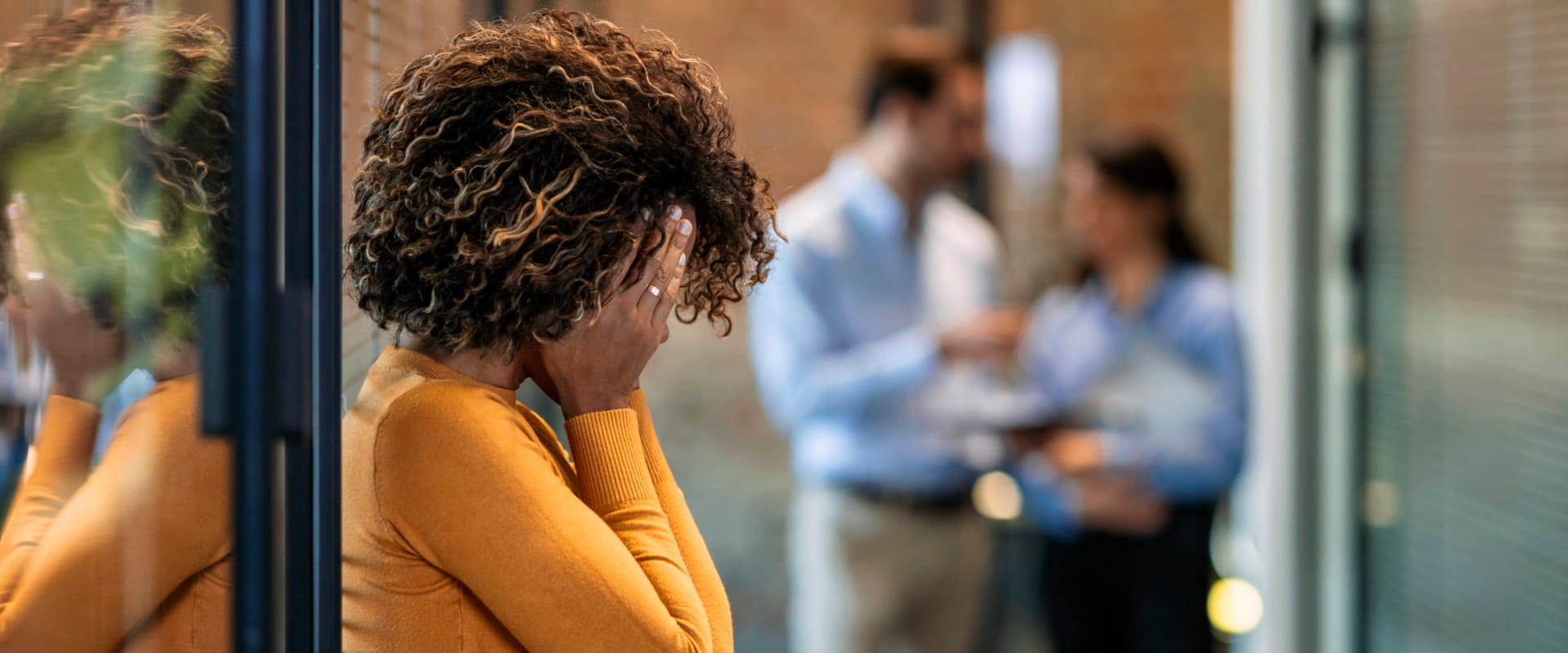 A person in an office stands in the hallway. Their hands are covering their face.