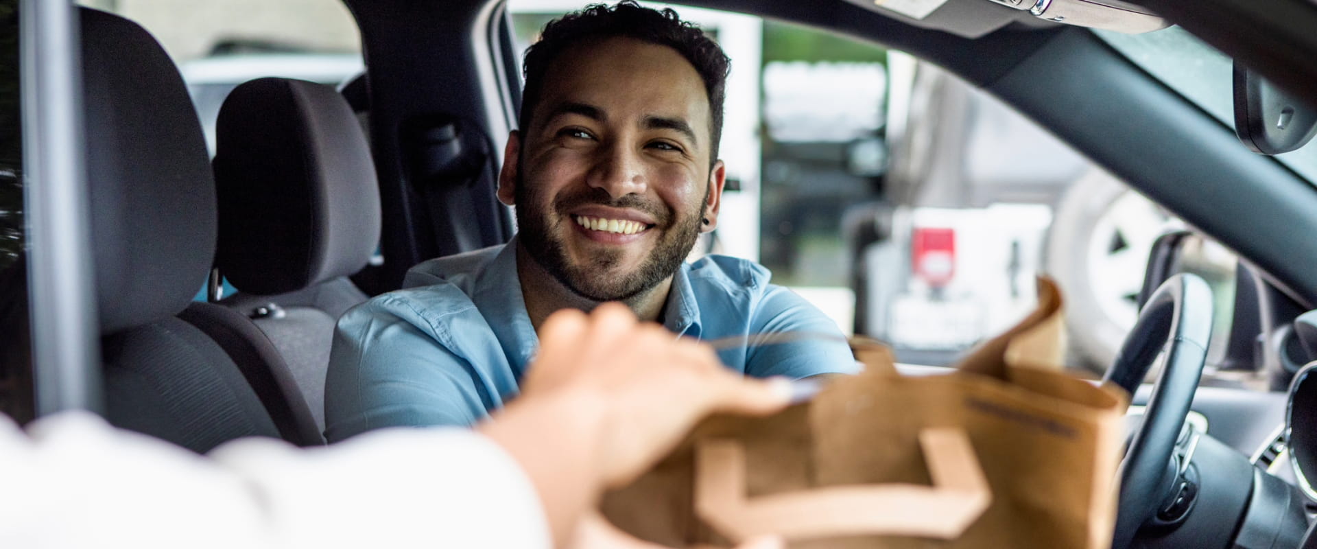 A smiling person sitting in the driver's seat of their car, reaches for a brown paper being handed to them from someone outside of frame"