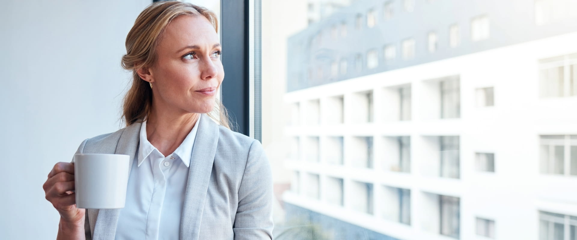 A business person looks out the window, holding a cup of coffee and their cell phone