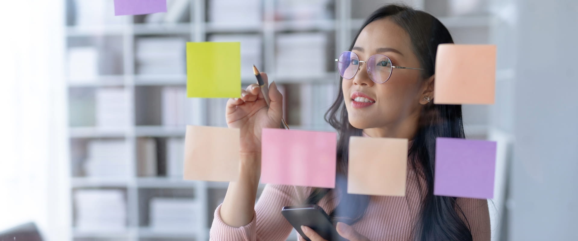 Professional individual writing on sticky notes and holding smartphone.