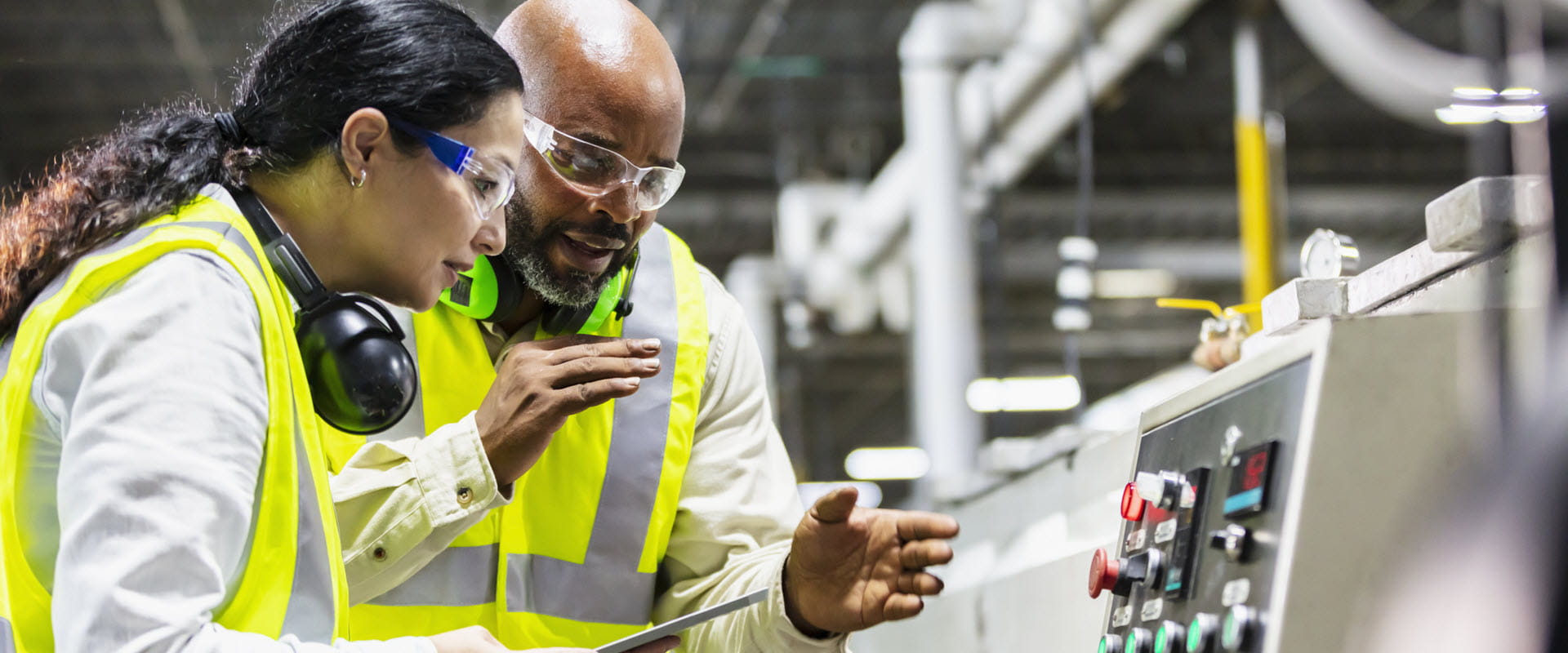 Two workers are in yellow safety vests and safety glasses. One worker is explaining something to the other while they look at a piece of equipment. Two workers are in yellow safety vests and safety glasses. One worker is explaining something to the other while they look at a piece of equipment.