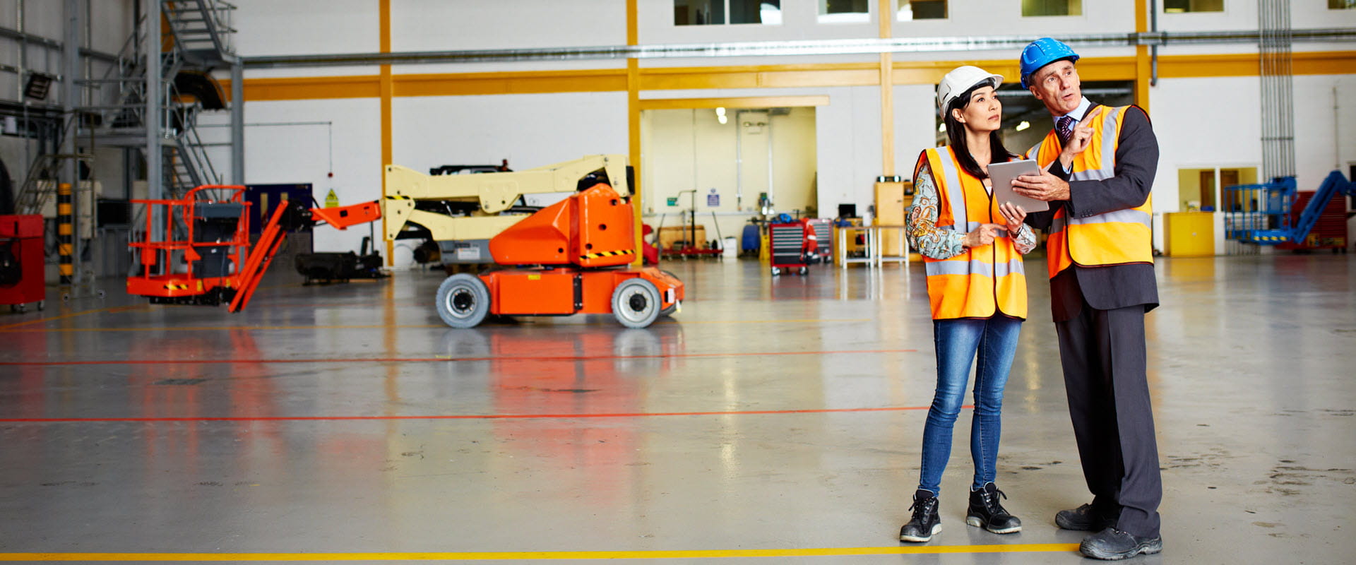 An individual, in a suit, hard hat, and safety vest, shows their tablet to another person, in jeans, a hard hat, and safety vest, in an empty warehouse An individual, in a suit, hard hat, and safety vest, shows their tablet to another person, in jeans, a hard hat, and safety vest, in an empty warehouse
