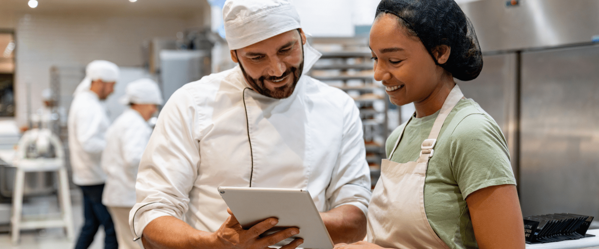Two workers in hair nets and aprons talk to each other while looking at a tablet. They are in a commercial kitchen.