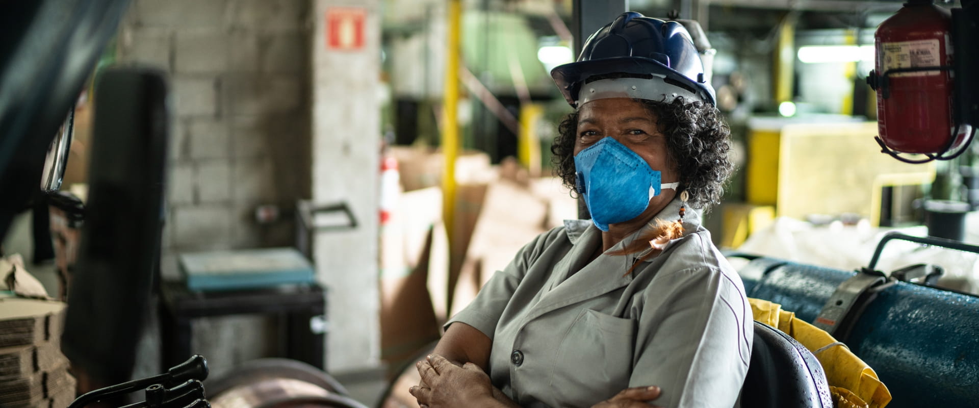 An older worker sits in a forklift, looking at the camera and wearing a mask An older worker sits in a forklift, looking at the camera and wearing a mask