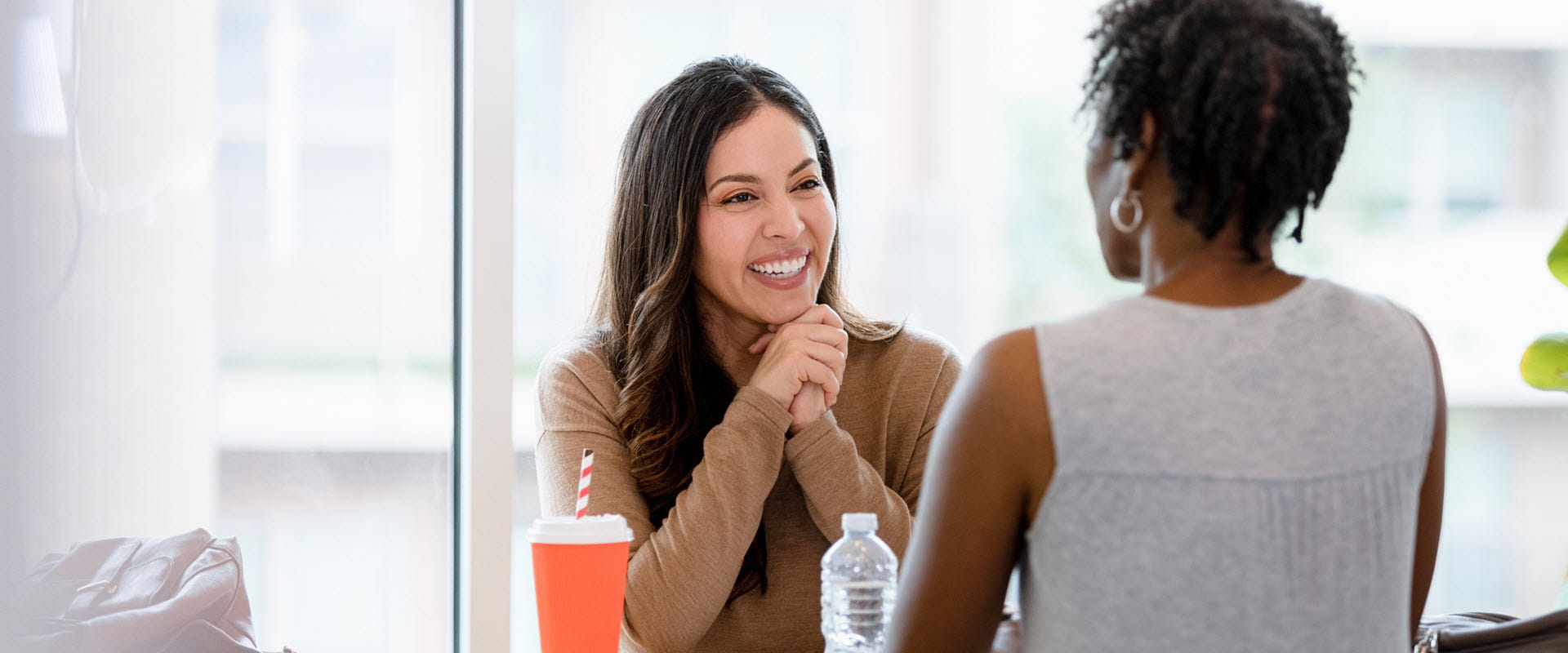 A smiling manager and employee sit across the table from one another in an office building. The table is covered in papers, coffee cup and water bottle. A smiling manager and employee sit across the table from one another in an office building. The table is covered in papers, coffee cup and water bottle.