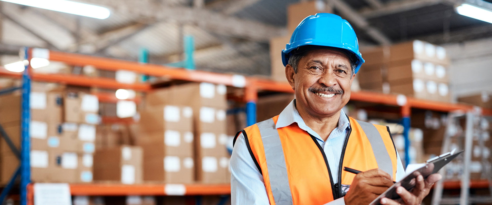 A worker in a warehouse wearing a blue hard hat and an orange safety vest is holding a clipboard.