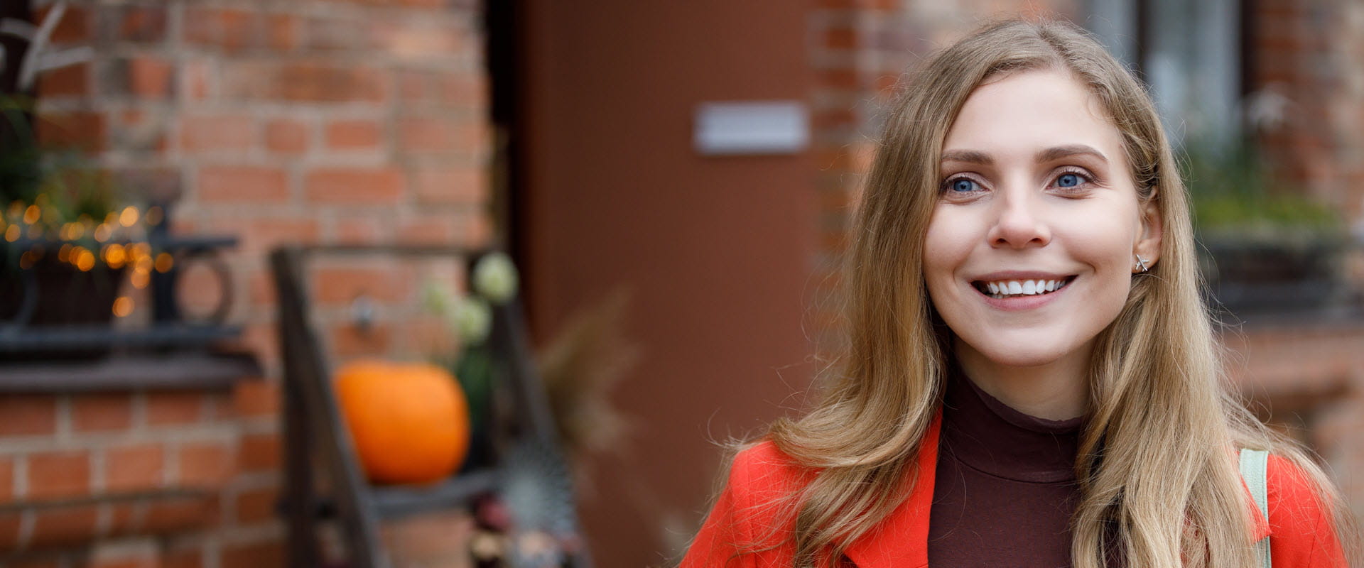 A smiling person standing in front of a brick building with autumn decorations.