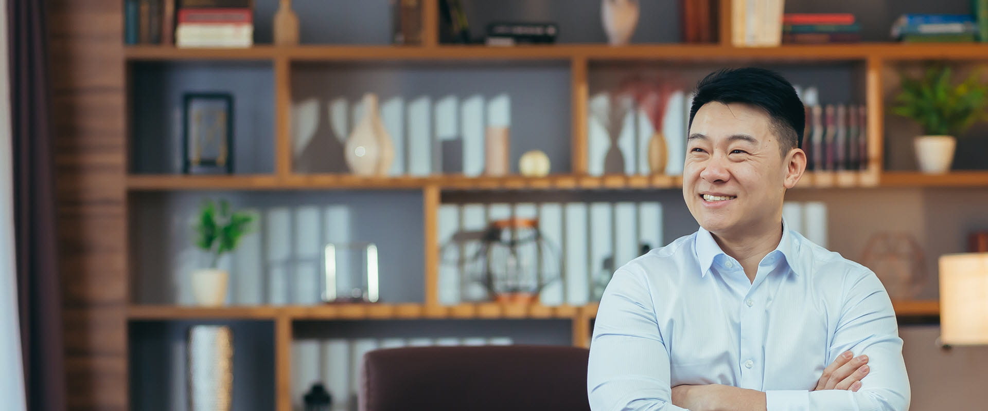 Smiling professional with arms crossed standing in a modern office with bookshelves in the background.