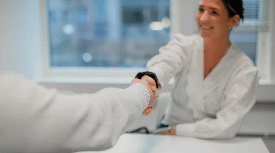 Kvinne smiler mens hun tar håndtrykk med en person under et møte på et kontor. Kvinne smiler mens hun tar håndtrykk med en person under et møte på et kontor.
