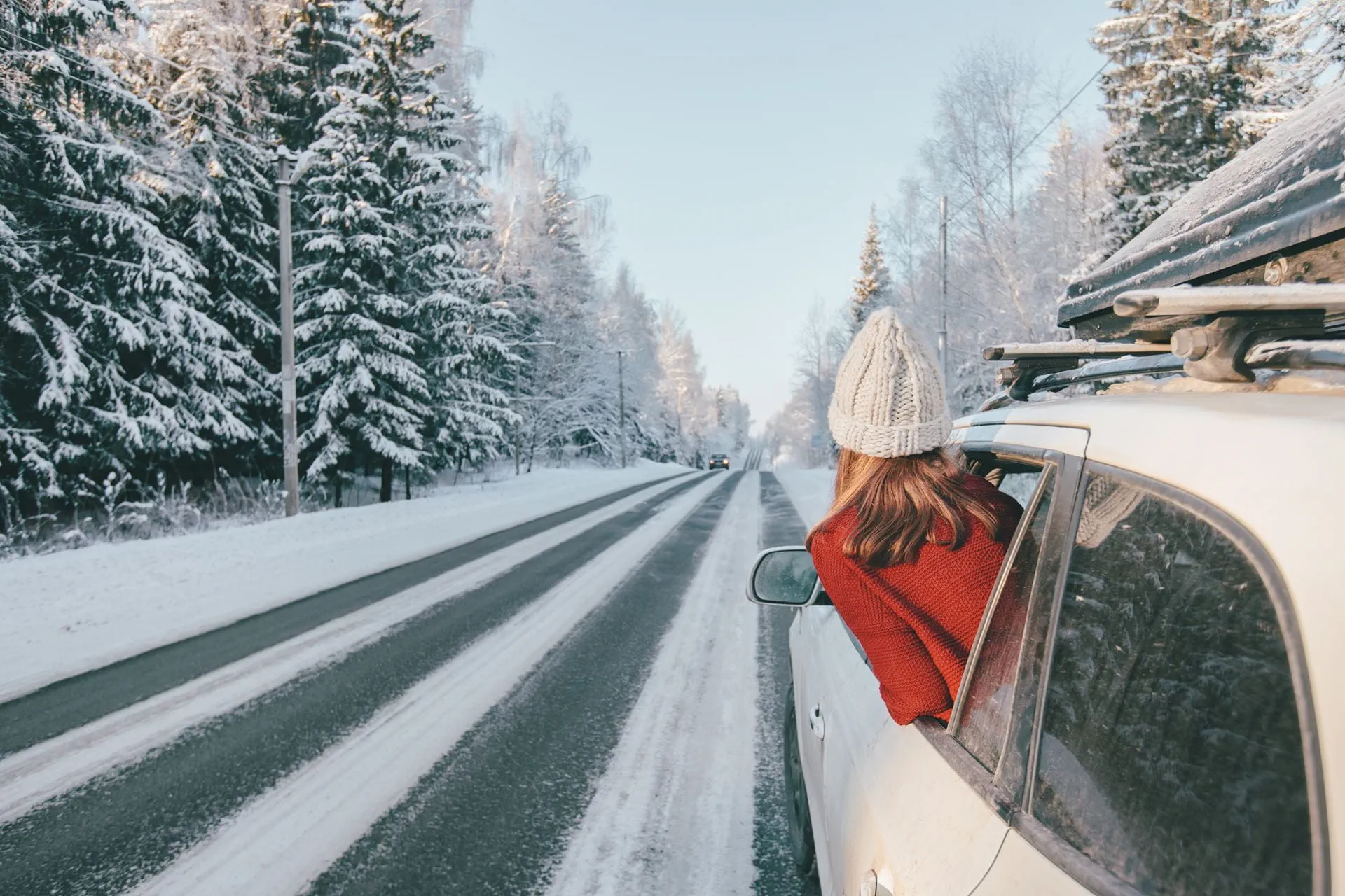 A woman wearing a red sweater and a  white knitted hat leans out of a car window,  enjoying the view of a scenic winter road  lined with snow-covered trees. The road  stretches into the distance under a clear  blue sky, creating a peaceful and  adventurous atmosphere.
