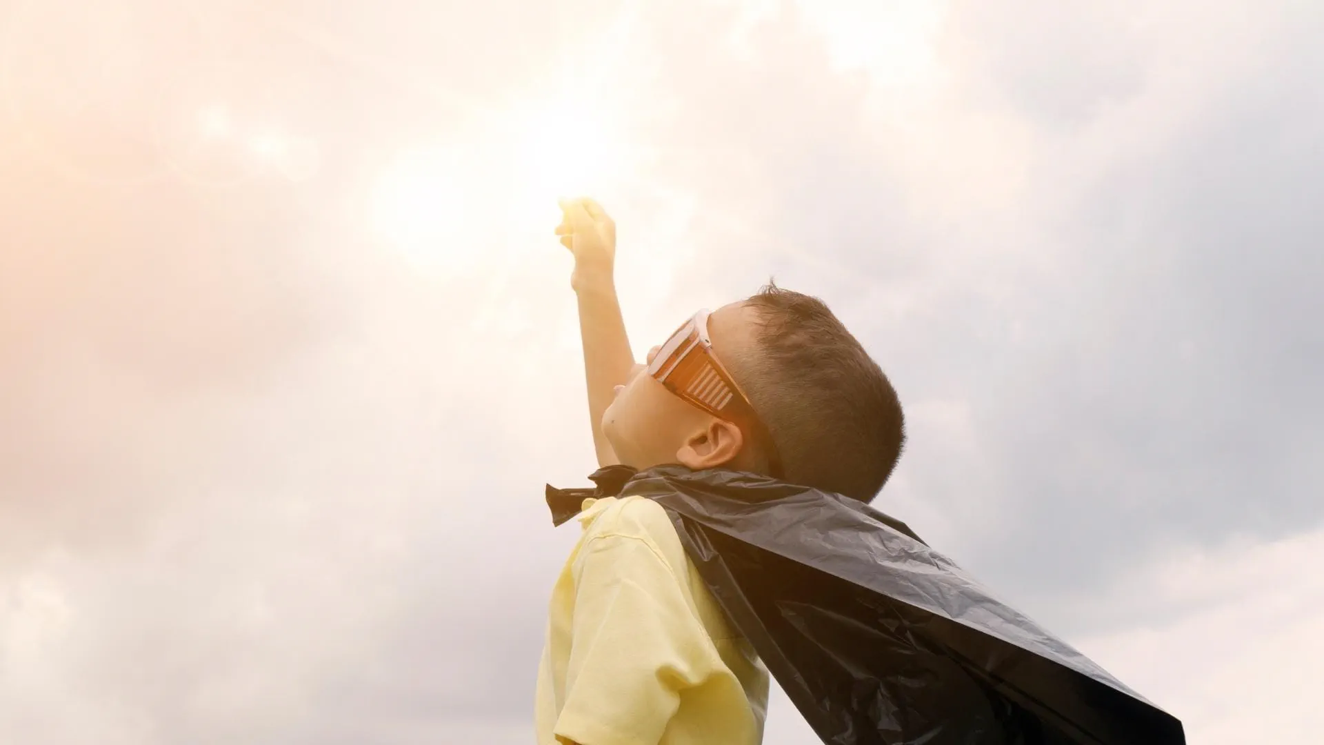 Ung gutt utkledd som superhelt med  solbriller og kappe strekker seg mot solen.  Inspirerende og drømmende stemning som  symboliserer ambisjoner, fantasi og  fremtidsmuligheter.