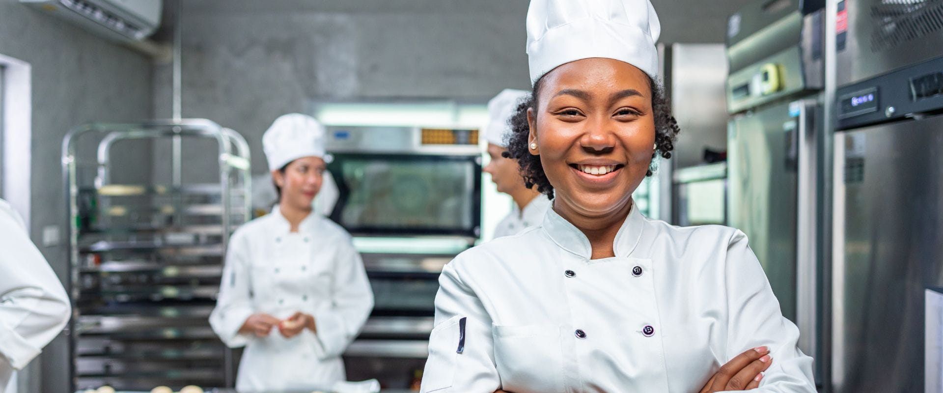 Smilende kokk i hvit uniform og lue  står i et profesjonelt kjøkken med  kollegaer i bakgrunnen. Moderne  kjøkkenutstyr og ovner viser et aktivt  og profesjonelt arbeidsmiljø innen  matlaging og gastronomi.