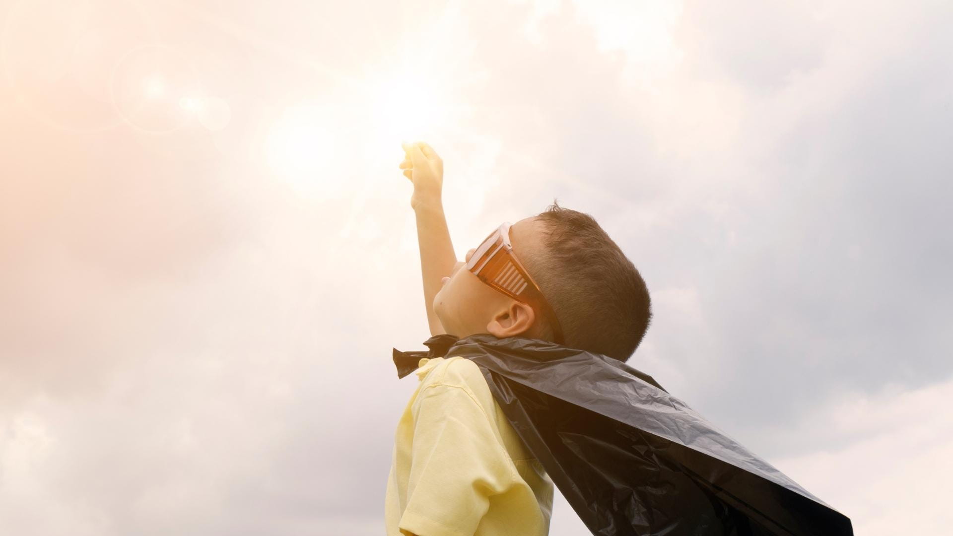 Ung gutt utkledd som superhelt med  solbriller og kappe strekker seg mot solen.  Inspirerende og drømmende stemning som  symboliserer ambisjoner, fantasi og  fremtidsmuligheter.