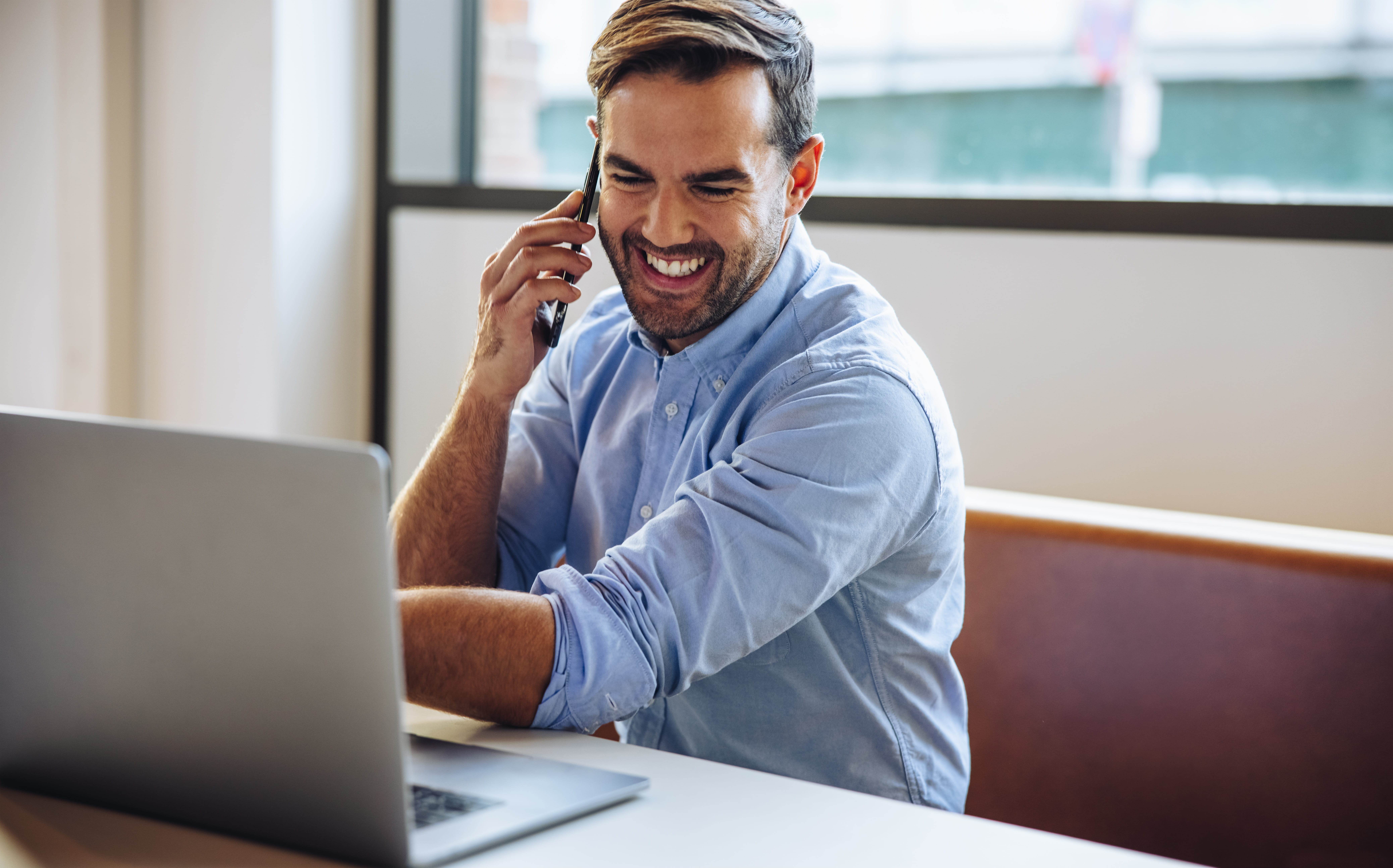 Smilende mann i blå skjorte sitter ved en  laptop og snakker i telefonen. Moderne  kontormiljø med naturlig lys fra store  vinduer. Produktivitet, profesjonelt arbeid  og digital kommunikasjon.