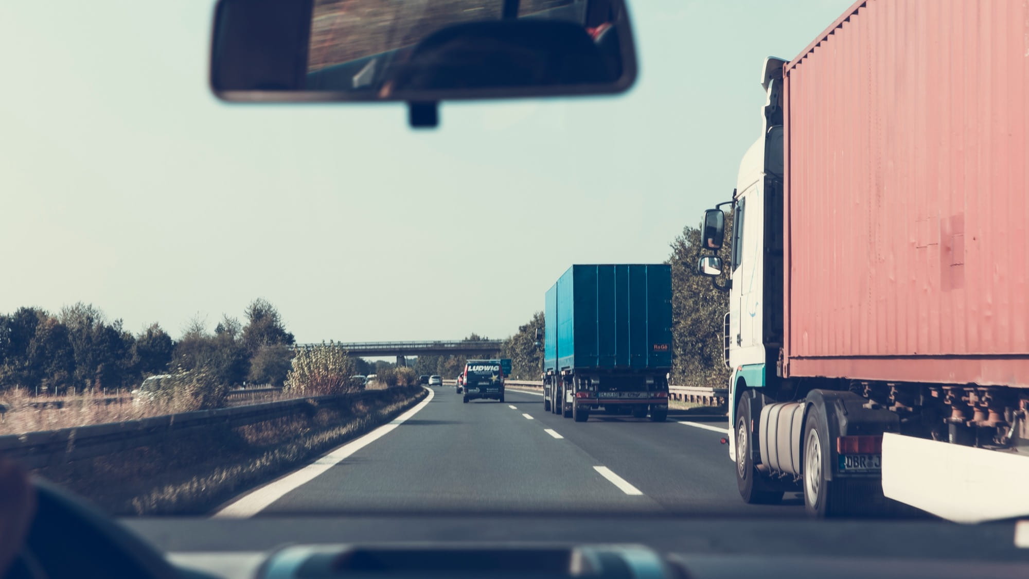 View from inside a vehicle driving on a  highway, surrounded by trucks and other  vehicles. The side mirror reflects the road  behind, while trees and a bridge line the  horizon.