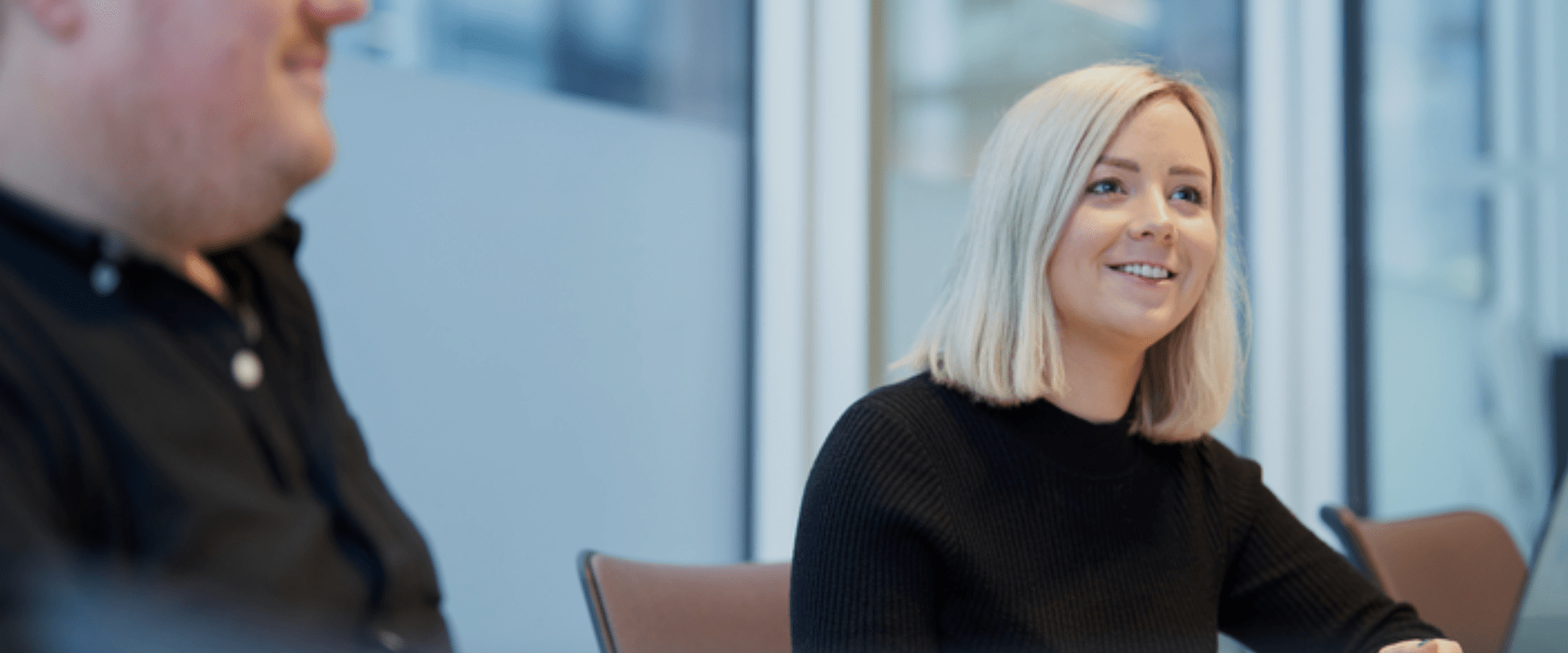 Smilende kvinne med blondt hår deltar i et møte i et moderne kontormiljø. En kollega i svart skjorte sitter i bakgrunnen. Profesjonell og engasjert samtale i en lys og stilren arbeidsplass.