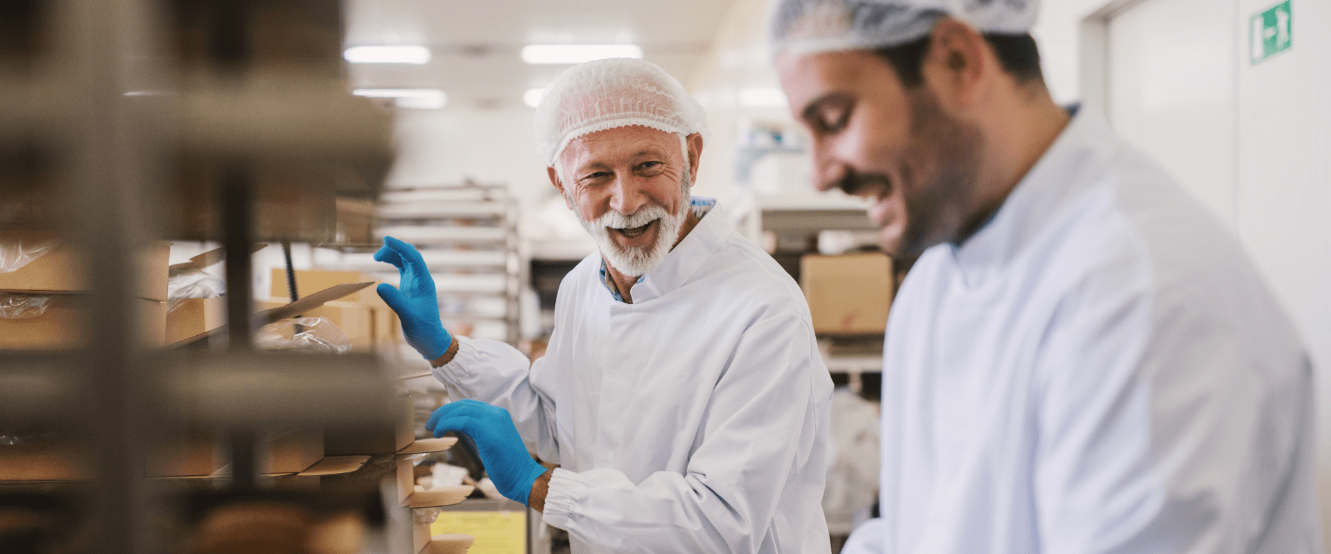Glade medarbeidere i produksjon,  iført hygieneklær og hårnett,  samarbeider på en matfabrikk.